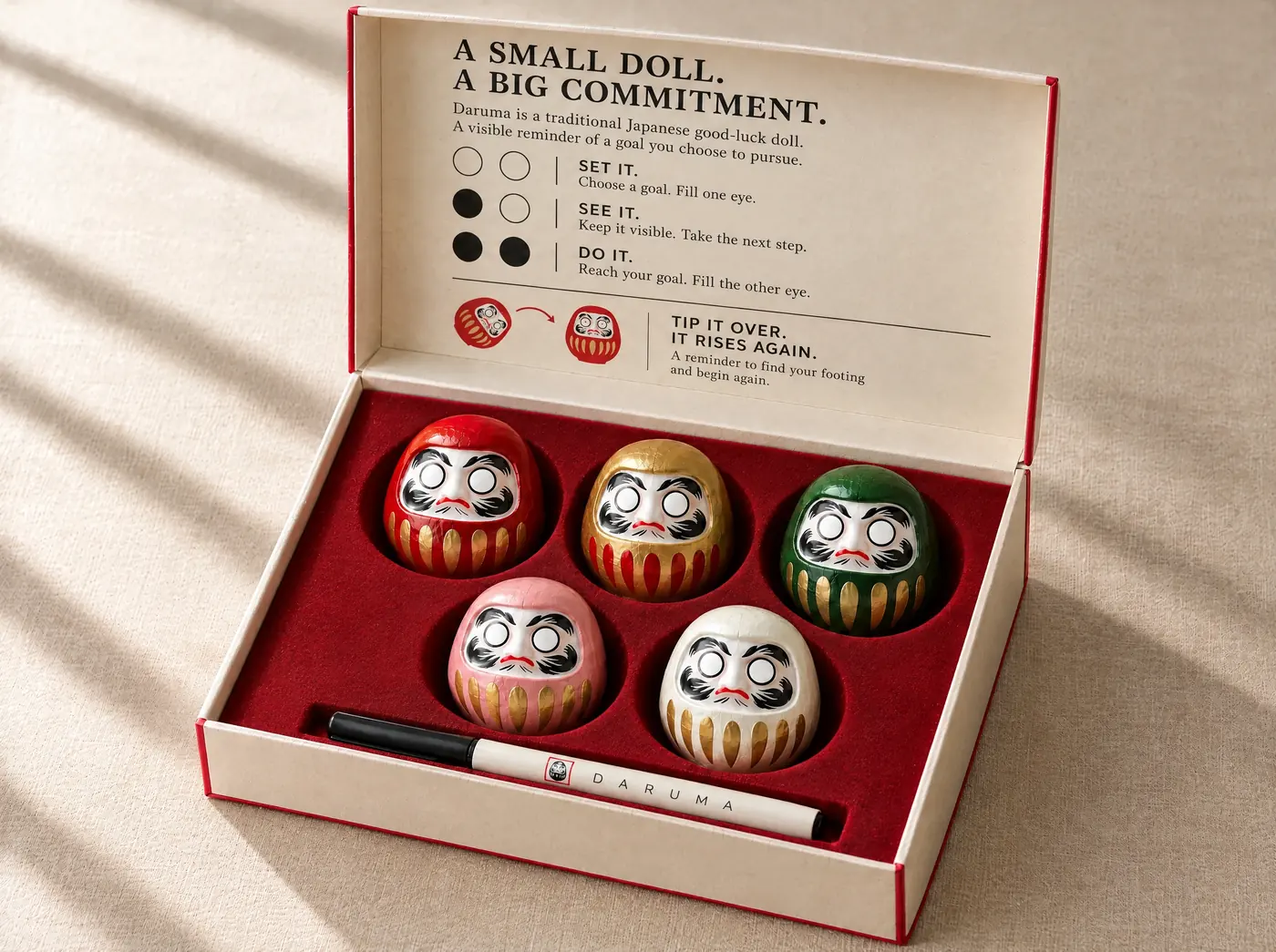An open gift box holding five Daruma dolls in red, gold, green, pink and white, with a marker pen in front.