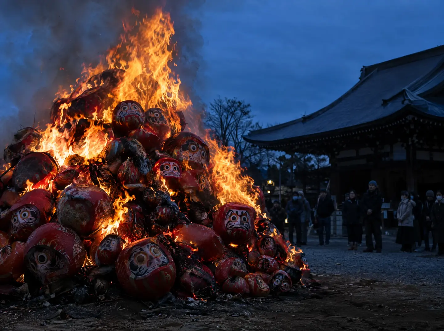 A large pile of old red Daruma dolls burns in a temple bonfire at dusk, with people watching quietly at a distance.